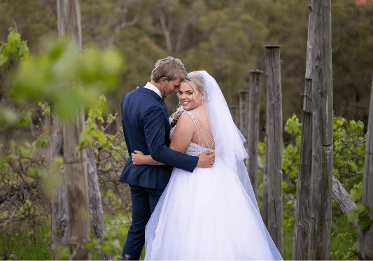 1 bride looking at photographer while holding groom in vineyards western australia